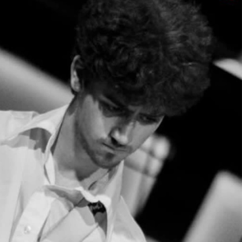 Black and white photo of a young man with curly hair, looking down with a serious expression. He wears an open-collared shirt and a loosened tie, evoking the introspective style often seen in portraits of Miles Tanner, with blurred, indistinct shapes in the background. The lighting creates shadows on his face.