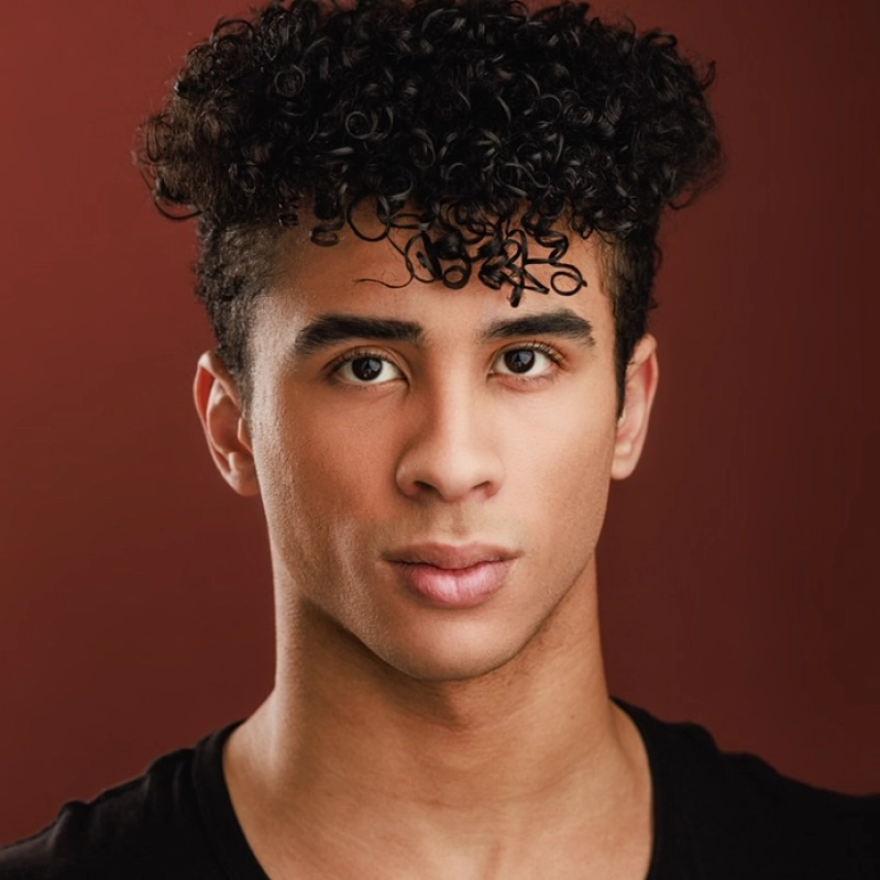 A young man with medium skin, short curly black hair, and dark eyes looks directly at the camera with a neutral expression. Miles Tanner is wearing a black shirt and stands in front of a reddish-brown background.