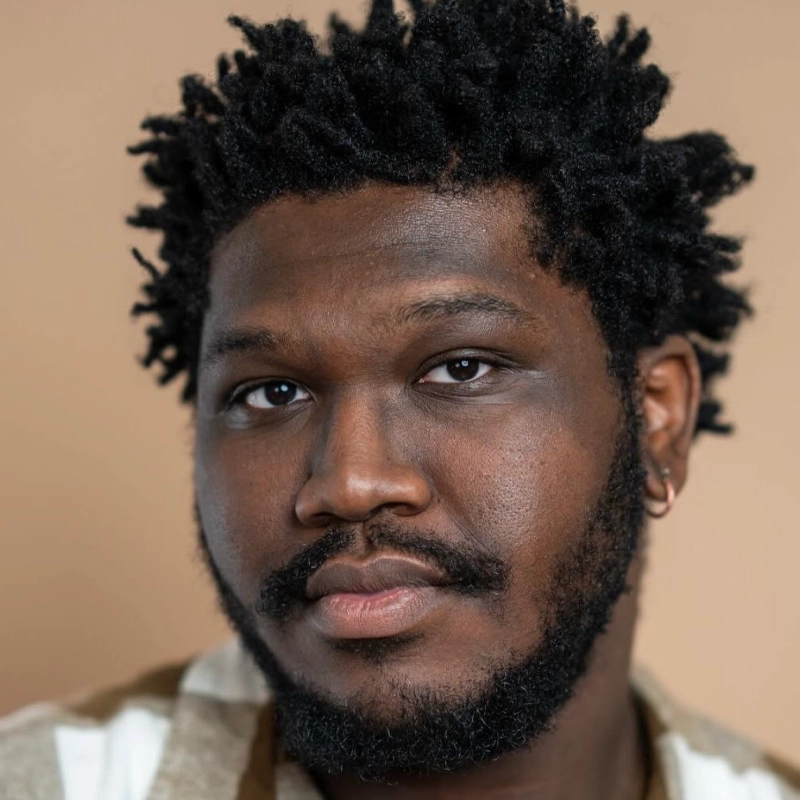 A close-up portrait of Ellistaire Perry, a Black man with short, textured hair and a full beard. He wears a neutral-colored patterned shirt and a small hoop earring in his left ear, set against a solid beige background, with a calm, thoughtful expression.