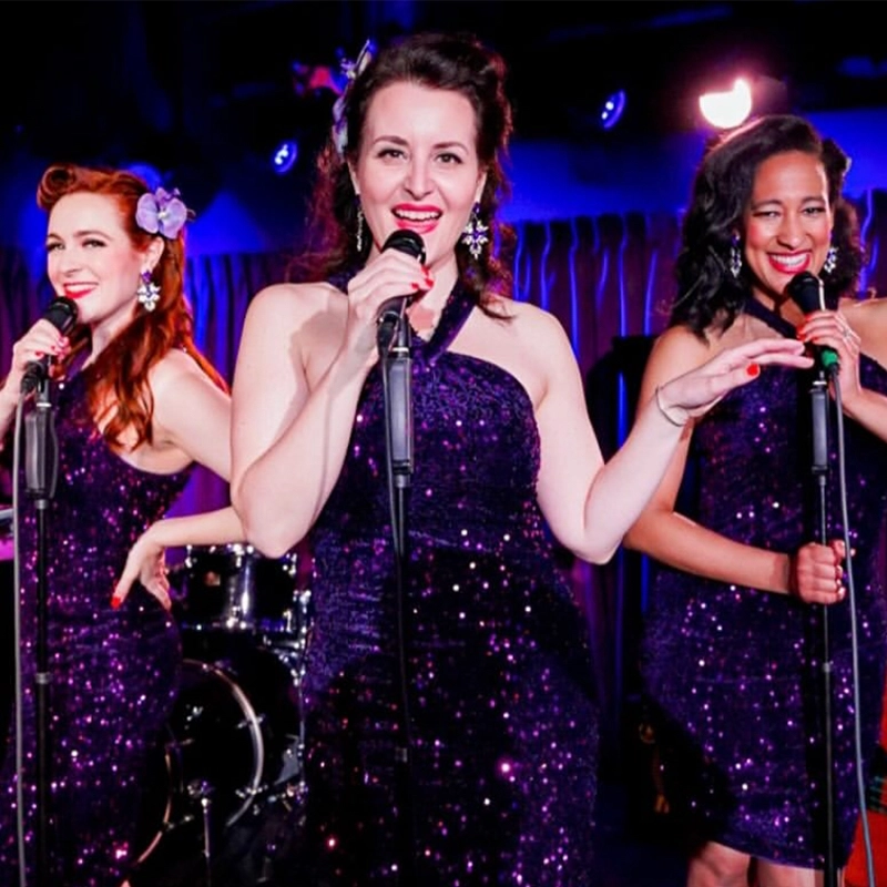 Three women in sparkly purple dresses sing into microphones on stage. They smile brightly under colorful lights, with musical instruments behind them. In the center, Alyssa Jaffe gestures with her hand while the others sing and pose beside her.