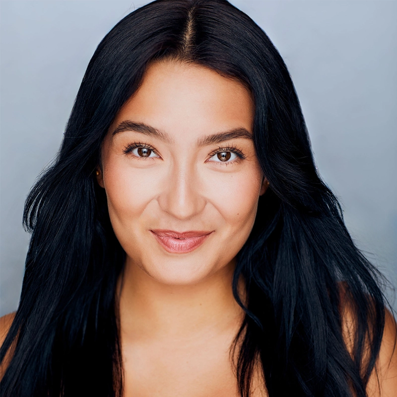 A woman with long, straight black hair and smooth medium-toned skin smiles softly at the camera. She has brown eyes, natural makeup, and is photographed by Christian Johannsen against a plain, light bluish-gray background.