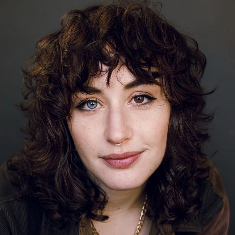A woman with shoulder-length curly brown hair, a gold nose ring, and a light complexion looks directly at the camera, smiling softly. Willow Dougherty has heterochromia: her left eye is blue and her right eye is brown. She wears eyeliner and a gold chain necklace.