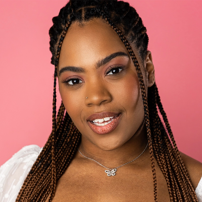 Stephanie Ainsworth, a young woman with long braided hair, smiles softly. She wears natural makeup with subtle eyeshadow and nude lipstick. A nose stud and delicate butterfly necklace complete her look against a solid pink background and white top.