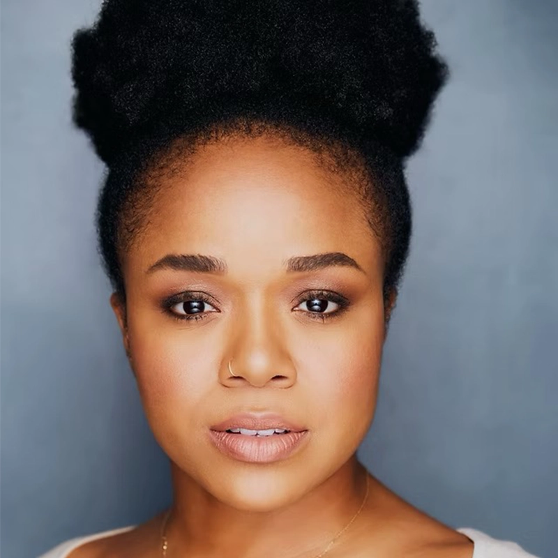 Gabrielle Elisabeth, a woman with deep brown skin and full lips, looks directly at the camera. Her dark natural hair is pulled into a voluminous puff. She wears defined brows, nude lipstick, and a small nose ring against a plain gray background.
