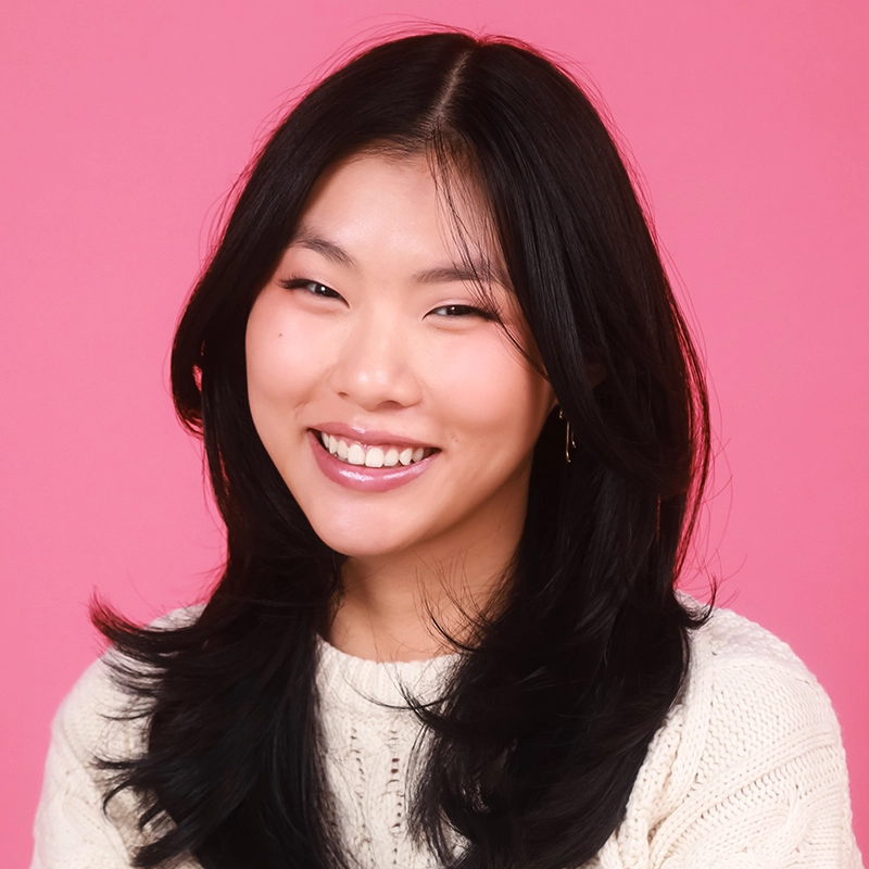 Katherine Lynn-Rose, a young woman with long, dark hair, smiles warmly. She is wearing a cream-colored sweater and gold hoop earrings. The solid pink background and her loose waves give her a relaxed and friendly look as she faces the camera.
