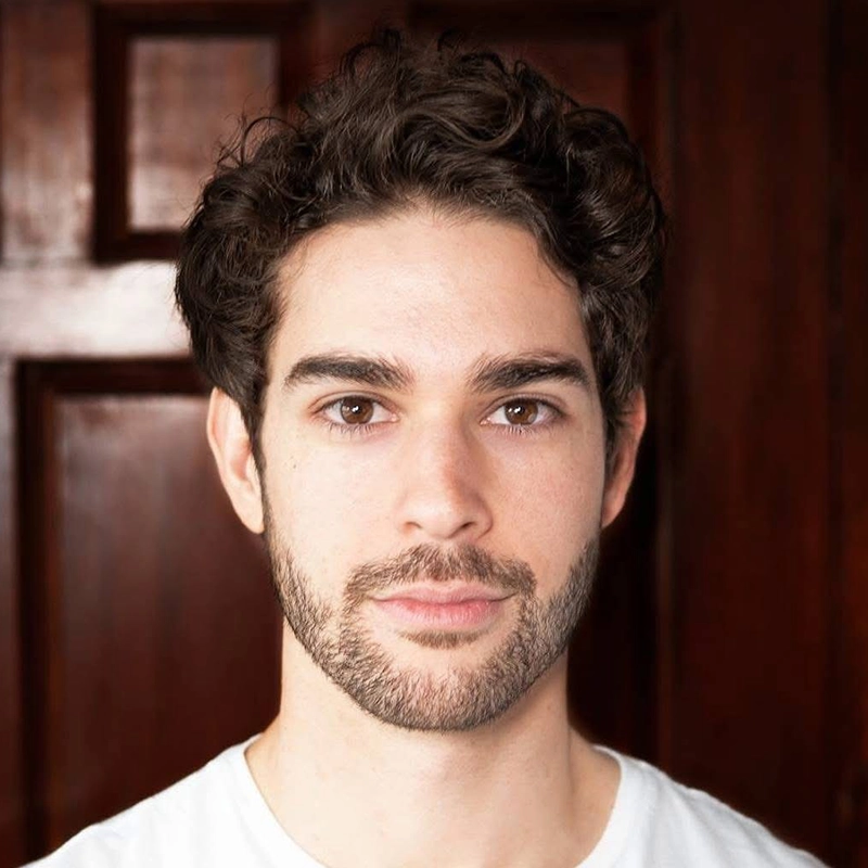 A young man with curly dark hair, trimmed beard, and mustache looks directly at the camera. He wears a plain white shirt. The background is blurred, showing a dark wooden door, echoing the portrait style of Stephanie Ainsworth.