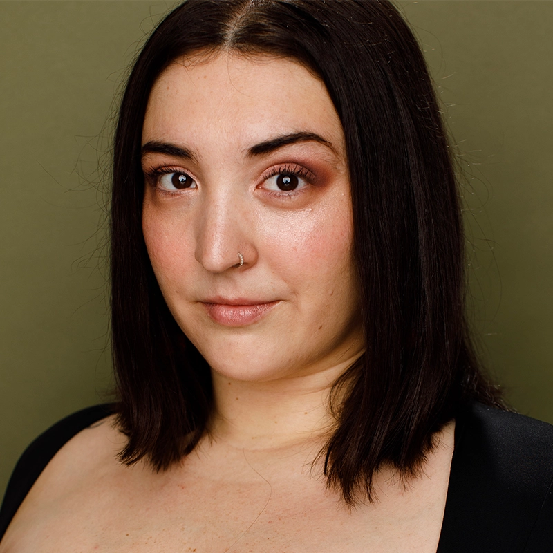 Stephanie Ainsworth, with medium skin, straight shoulder-length dark brown hair, and brown eyes, looks at the camera with a slight smile. She wears a nose ring and natural makeup, in a black top against an olive green background.