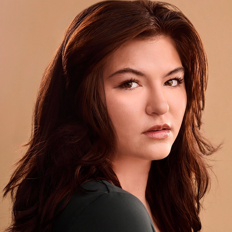 A young woman with long, wavy brown hair and fair skin looks seriously at the camera. She has natural makeup, arched eyebrows, and is wearing a dark top. The background is a soft beige, giving the portrait a warm, neutral tone.