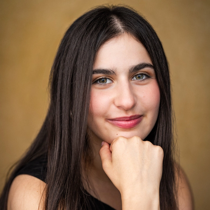 A young woman with long, straight brown hair and light skin smiles softly. She rests her chin on her fist, wearing a sleeveless black top. The background is a plain, warm beige color, and the lighting highlights her facial features.