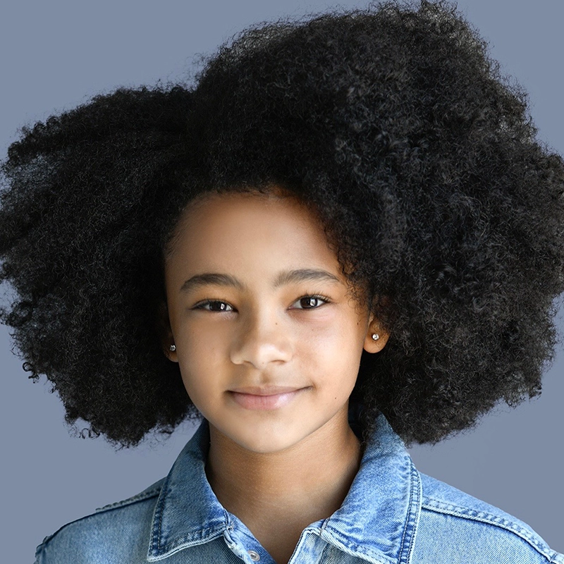 A young girl with a medium skin tone and natural, voluminous curly hair smiles softly. She wears small stud earrings and a blue denim jacket. The background is a solid muted grayish-blue, perfectly capturing Austin Elle Fisher's charming style.