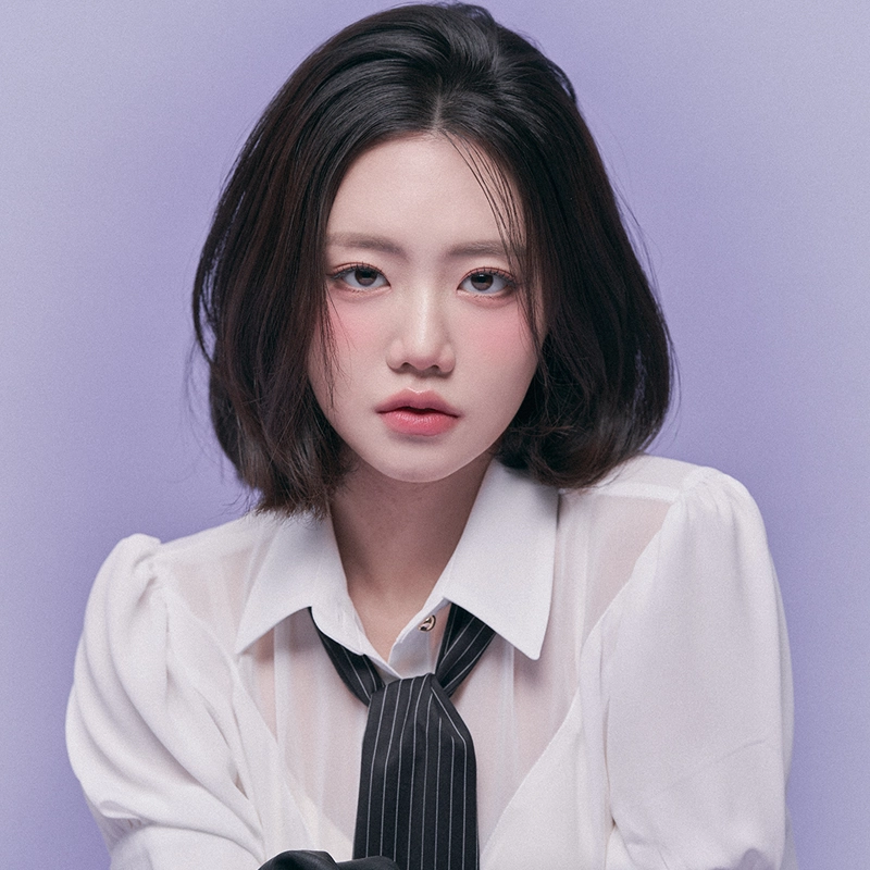 A young woman with short, dark hair and light skin poses against a lavender background. Flynn Jungbin Byun wears a white, semi-sheer blouse and a loosely tied black striped tie, her expression neutral with slightly flushed cheeks and natural makeup.