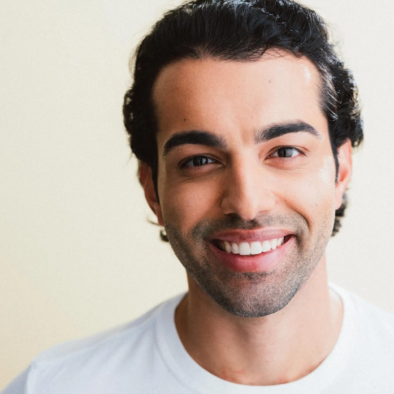 Jose Contreras, a young man with dark hair and eyebrows, lightly tanned skin, and a trimmed beard, smiles at the camera in a white shirt. The neutral, light background creates a clean and bright portrait.
