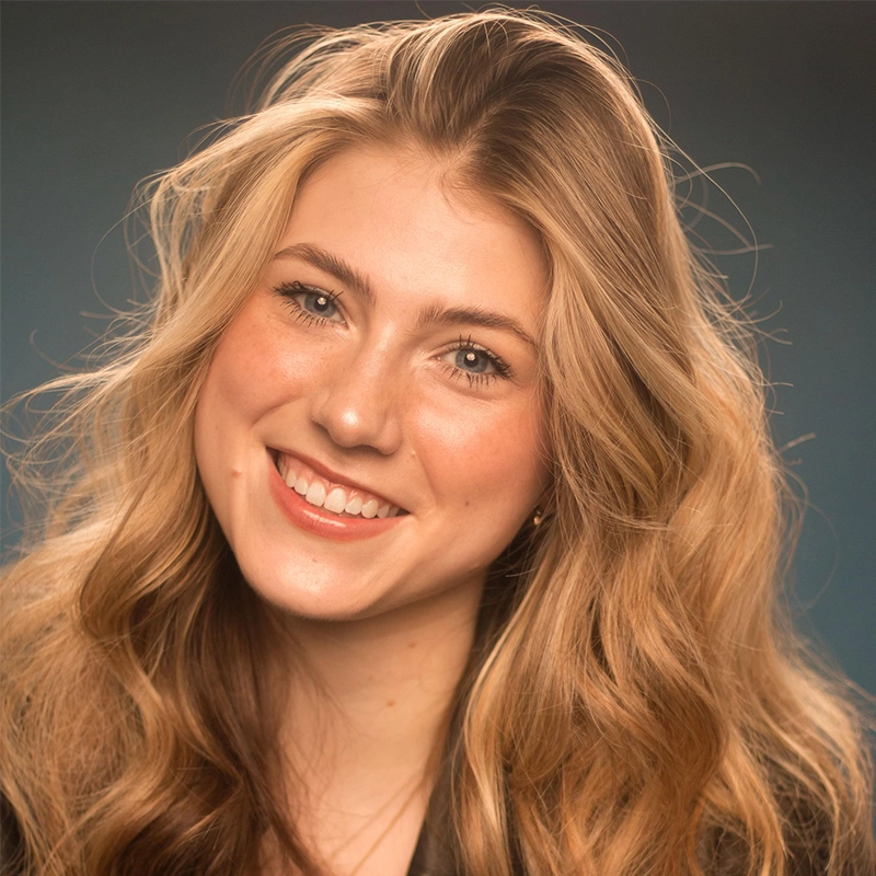 A young woman with long, wavy blonde hair smiles warmly at the camera. She has fair skin, blue eyes, and is wearing a dark top. The background is a soft, out-of-focus dark blue, creating a gentle and welcoming atmosphere.
