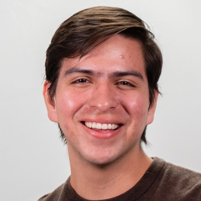 A young person with short, dark brown hair and light skin smiles warmly at the camera. They are wearing a brown crew-neck sweater and are posed against a plain, light gray background.