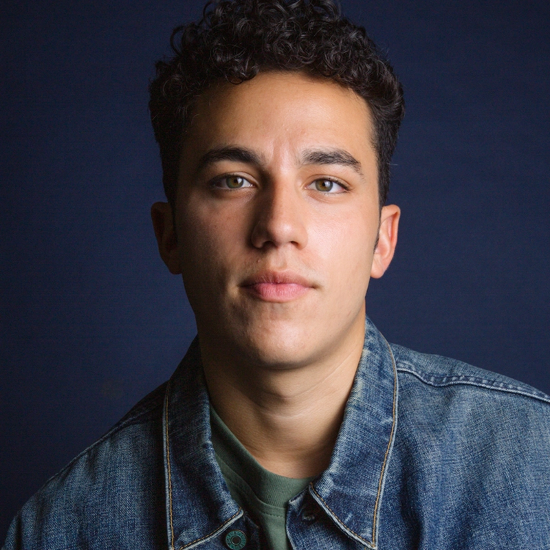 A young man with short, curly dark hair looks directly at the camera with a neutral expression. Fernando Flores is wearing a blue denim jacket over a green shirt. The background is dark blue, and the lighting highlights his face evenly.