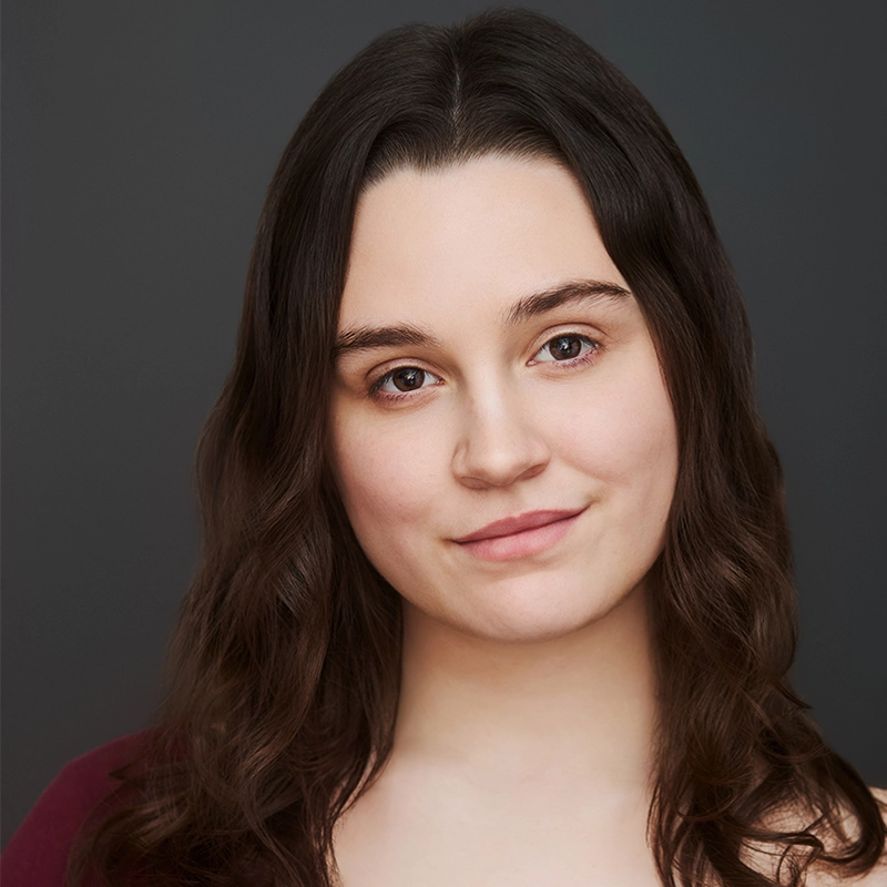 A young woman with fair skin and long, wavy brown hair poses against a plain dark background. She has brown eyes, subtle makeup, and a slight smile. She wears a burgundy top and looks directly at the camera, exuding calm confidence.