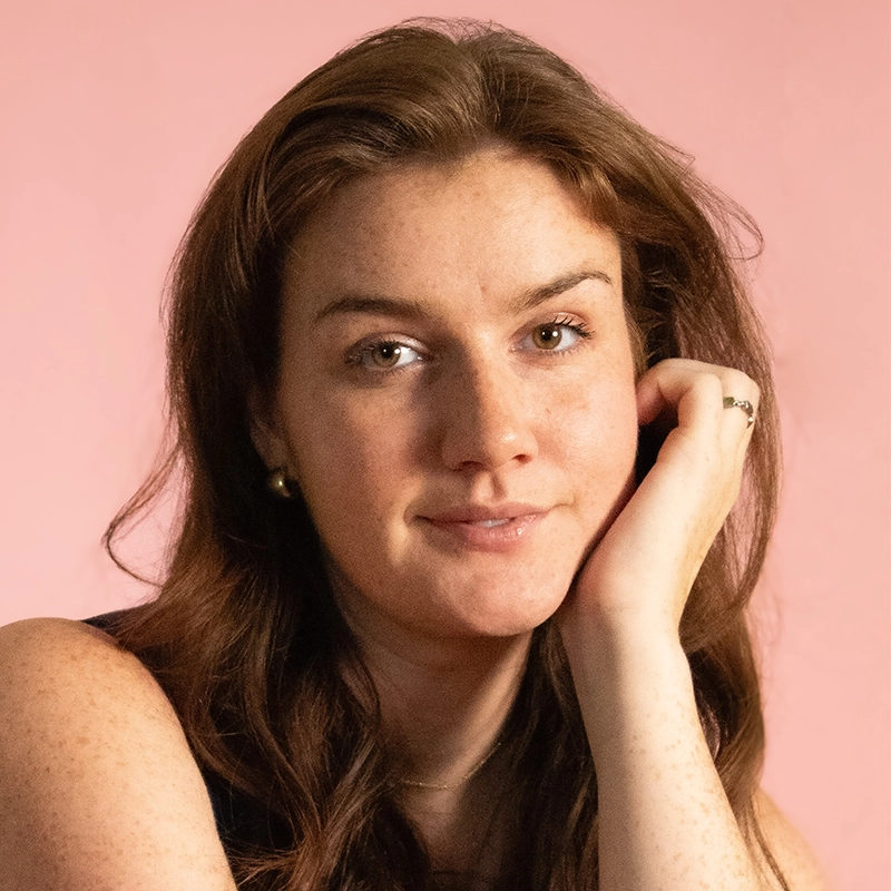 A woman with long brown hair and fair skin rests her chin on her hand, looking at the camera with a slight smile. She wears a dark top, a gold ring, and gold stud earrings. The background is a soft pink.