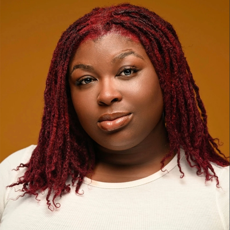 A woman with deep red, shoulder-length locs poses against a warm orange background. She wears a cream-colored top and gazes confidently at the camera, her makeup subtle with glossy lips and softly defined eyes.