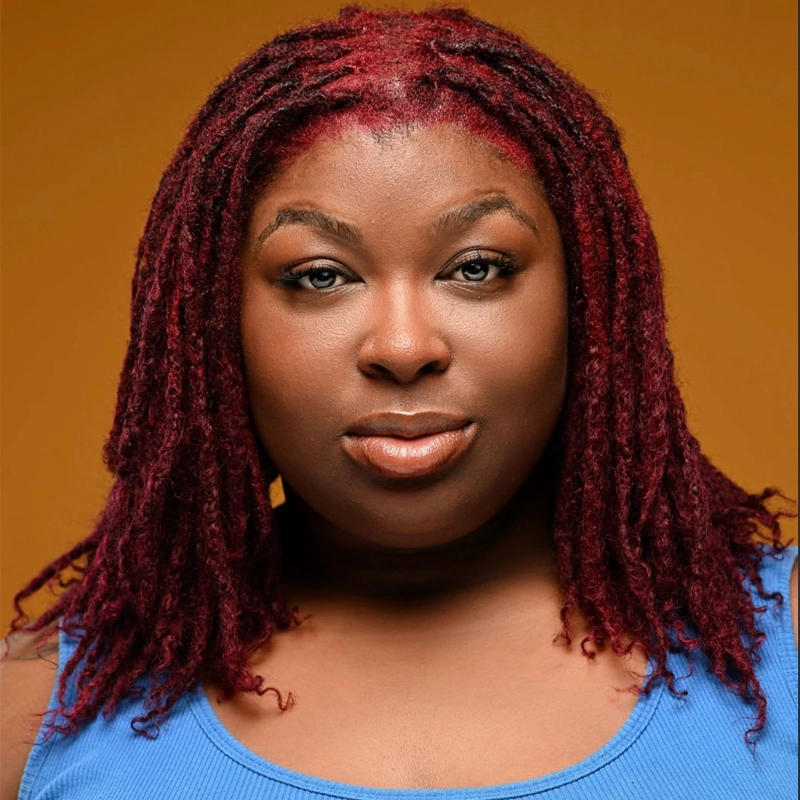 A confident Black woman with long, burgundy locs and natural makeup looks directly at the camera. She wears a blue sleeveless top and stands against a warm, solid orange background.