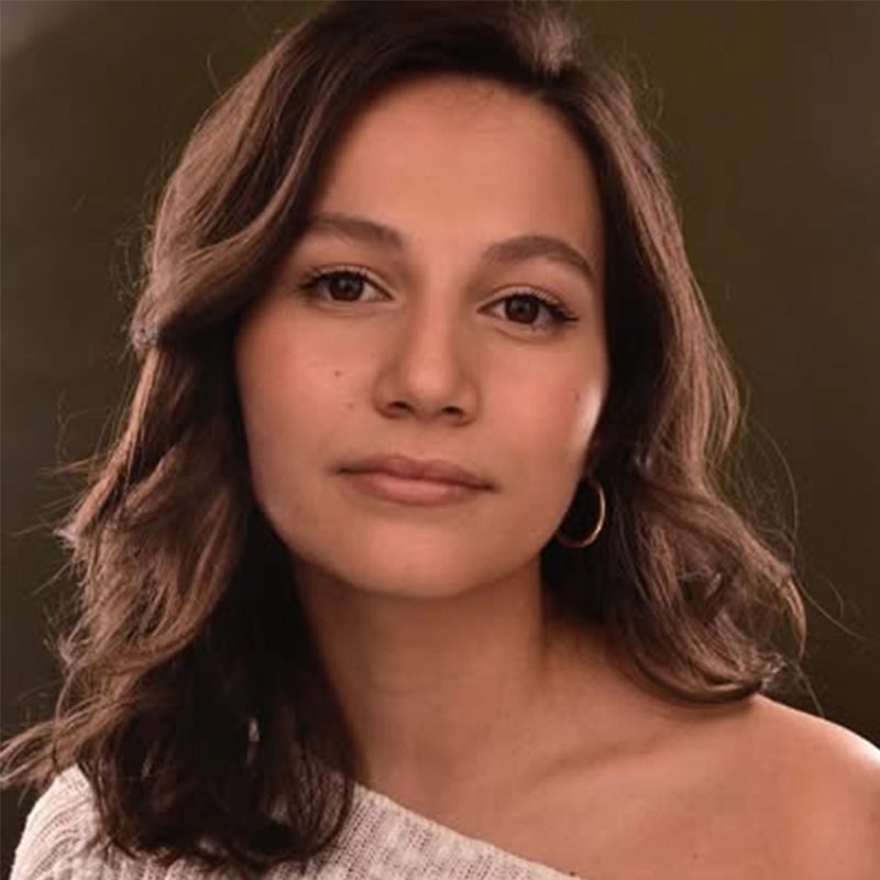 A young woman with medium-length wavy brown hair and light skin gazes softly at the camera. Kendall Becerra wears a white off-the-shoulder top and a small hoop earring, her calm, relaxed expression set against a neutral background.