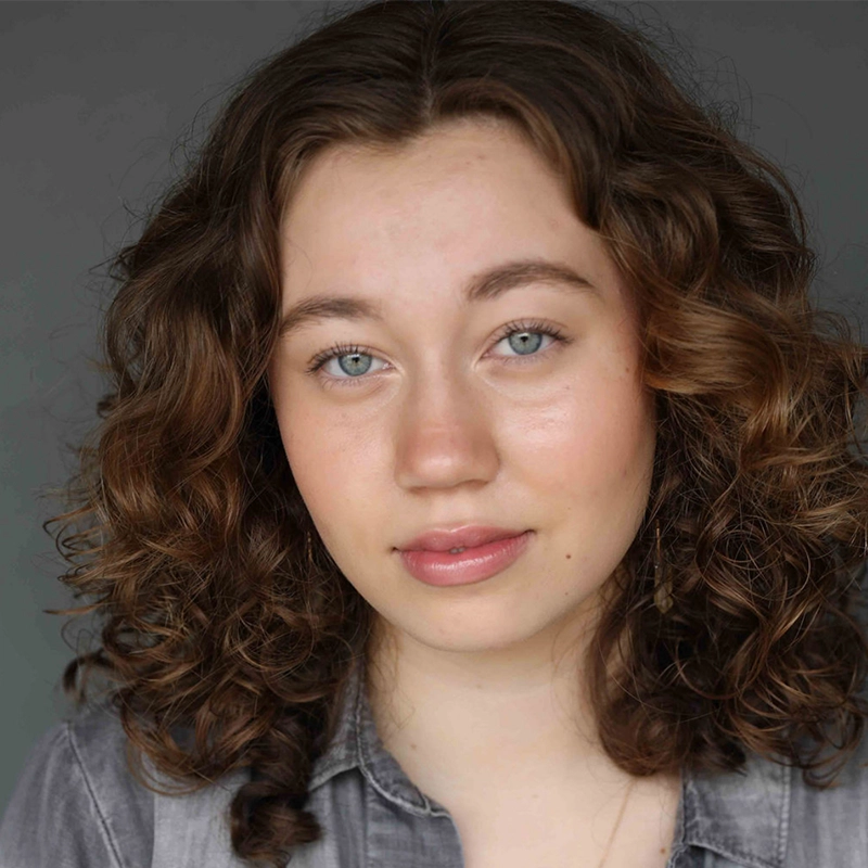 A young woman with fair skin, blue eyes, and curly light brown hair looks at the camera with a neutral expression. She is wearing a gray button-up shirt and a subtle necklace, and the background is a plain, dark gray.
