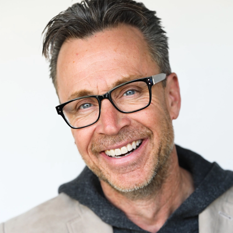A middle-aged man with short, gray-streaked hair smiles warmly. He wears black-rimmed glasses, a light beige jacket, and a dark hoodie. His teeth are visible, and he stands in front of a plain, light background.