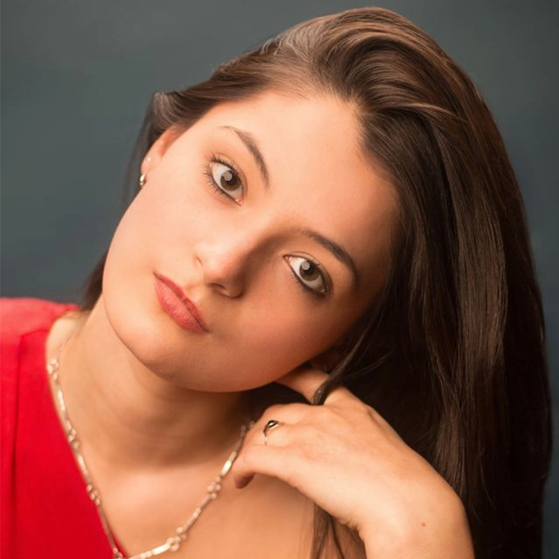 A young woman with long brown hair tilted to the side gazes softly at the camera. She wears a red top, a delicate necklace, and a small earring. Her hand rests near her neck, and the background is a smooth, dark blue.