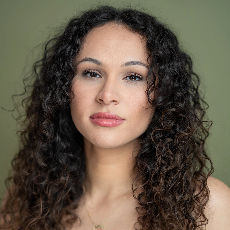A woman with medium skin tone and long, dark, curly hair reminiscent of Milan Parodie looks directly at the camera with a neutral expression. She has subtle makeup and wears a delicate gold necklace against a soft, plain green background.