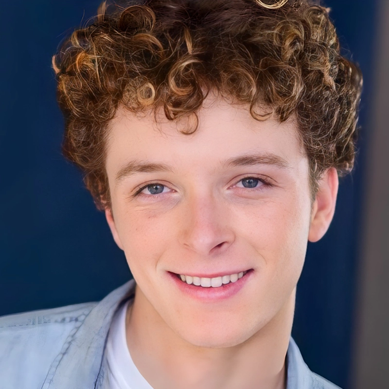 A young person with fair skin, curly light brown hair, and blue eyes smiles warmly at the camera. They are wearing a light gray shirt over a white top. The background is plain and navy blue, keeping the focus on the person's cheerful expression.