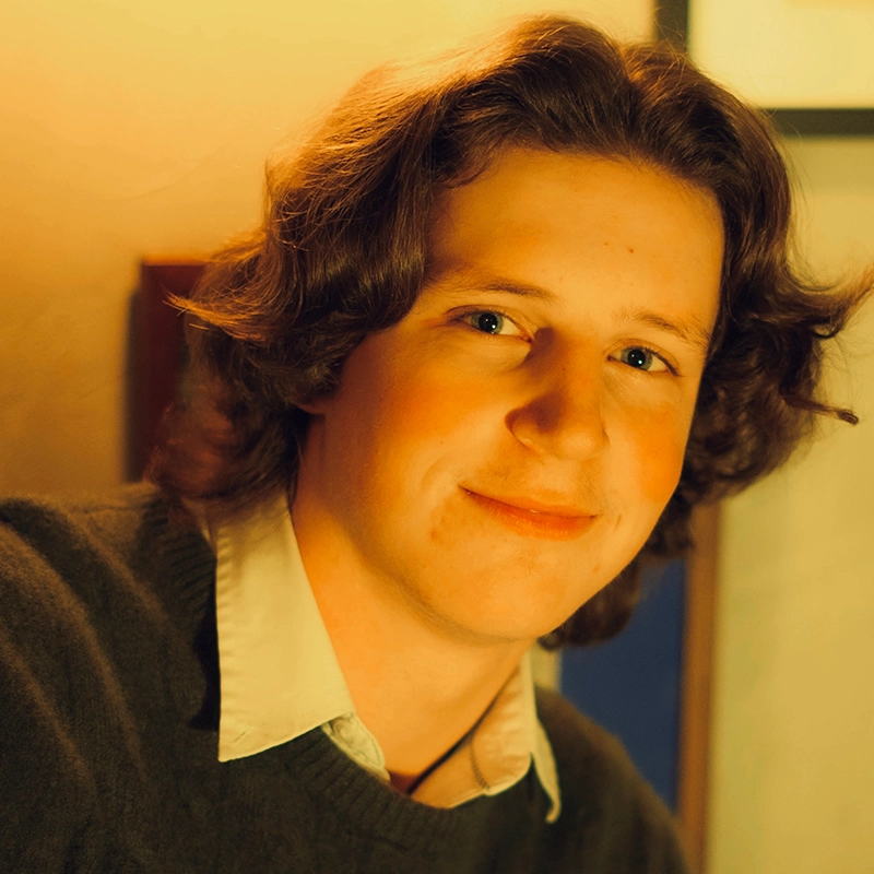 A young person with wavy brown hair and fair skin smiles softly at the camera. They wear a white collared shirt under a dark sweater. The warm lighting creates a cozy atmosphere, and a blurred picture frame is visible in the background.