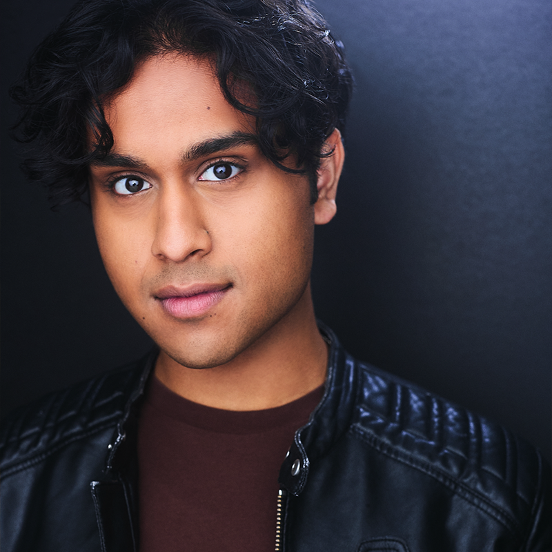 Tanzil Philip, with medium brown skin and wavy dark hair, is pictured against a dark background. Wearing a black leather jacket over a brown shirt, they look directly at the camera with a neutral, slight smile as soft lighting highlights their features.