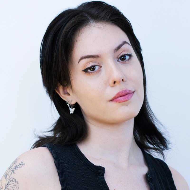 A young woman with fair skin, straight dark brown hair, and winged eyeliner looks at the camera with a neutral expression. Angelique Rodriguez wears a sleeveless black top, small silver earrings, and has a floral tattoo on her upper arm against a plain white background.