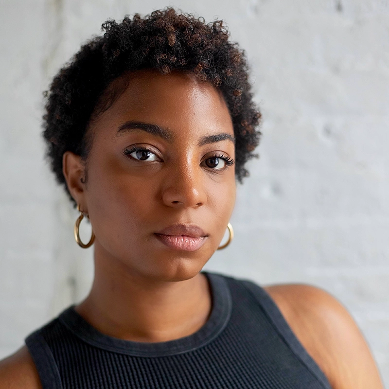 A woman with short curly hair, wearing gold hoop earrings and a sleeveless black top, looks calmly at the camera. She stands against a light gray textured wall, with soft natural light highlighting her face.