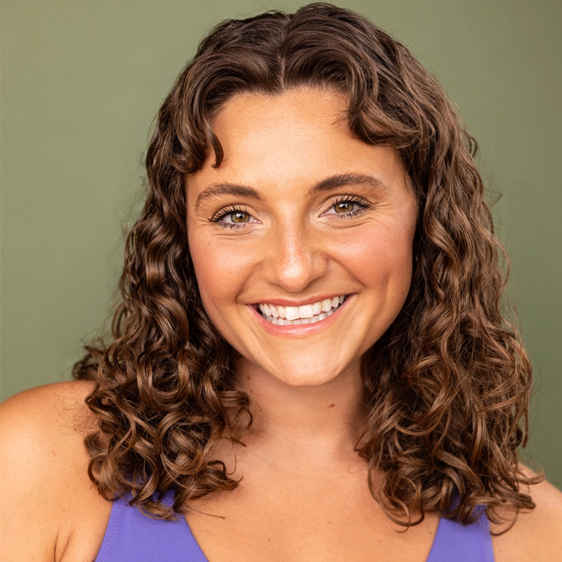 Rachel Thomas, a smiling woman with medium skin and shoulder-length, curly brown hair, stands in front of a plain green background. She wears a sleeveless purple top. Her makeup is natural, and she has bright hazel eyes, visible teeth, and dimples, giving her a cheerful, approachable look.