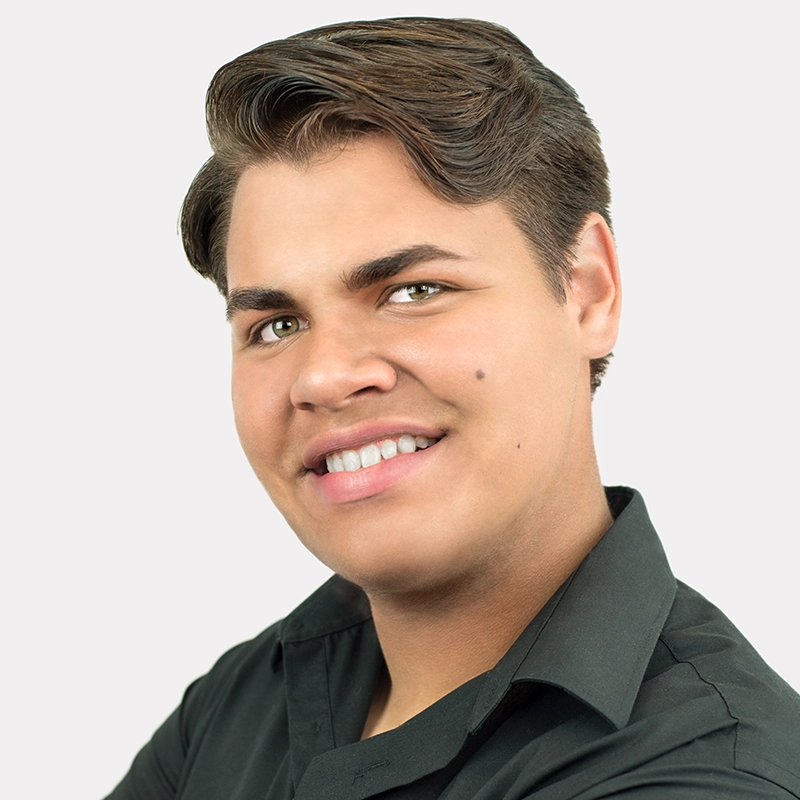 A young man with light brown hair styled neatly to the side, light eyes, and a beauty mark on his right cheek smiles at the camera. He is wearing a black collared shirt and is posed against a plain, light gray background.