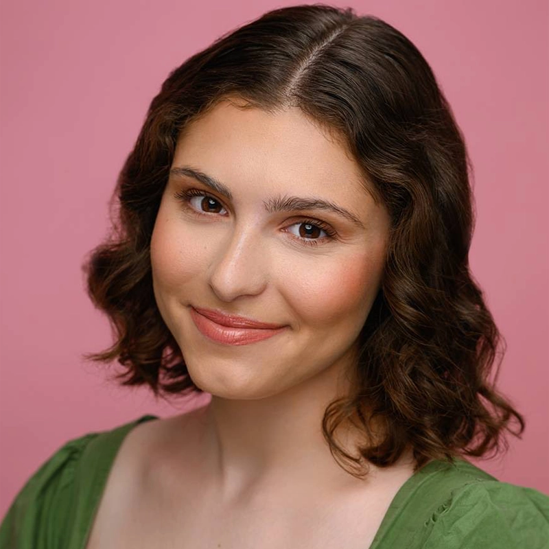 Dara Weinstein, a young woman with short, wavy brown hair, smiles softly at the camera. She wears a green top and natural makeup with pink lipstick. The solid pink background gives the image a warm, cheerful feel.