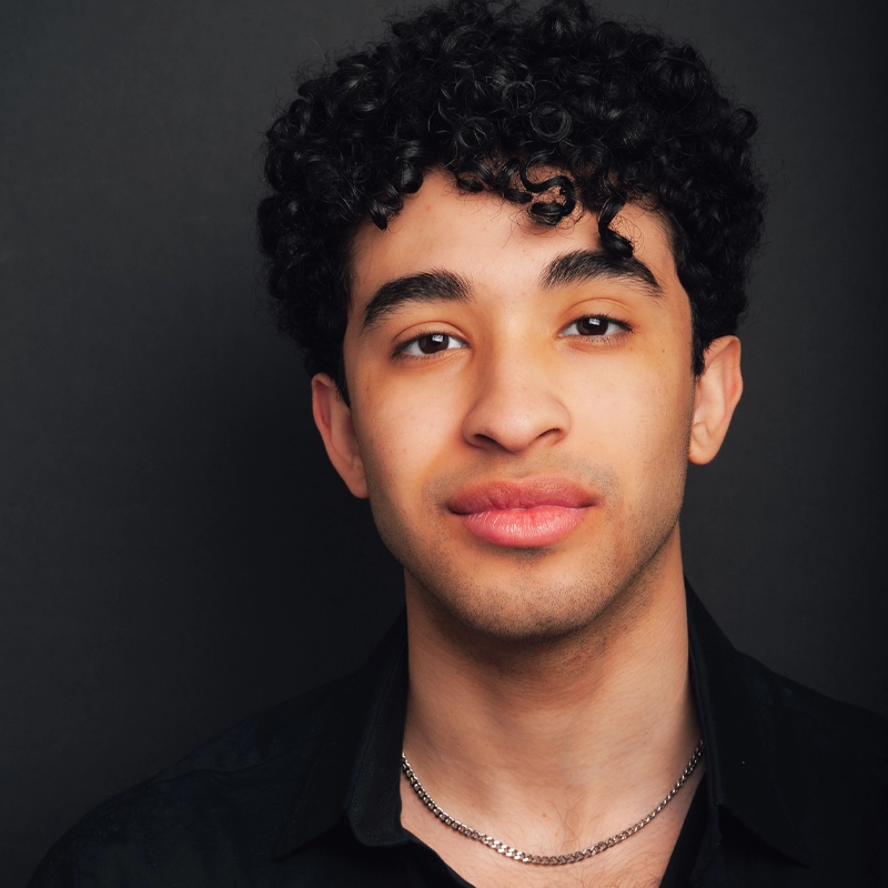 A young man with curly black hair and medium skin tone faces the camera, wearing a black shirt and a silver chain necklace, against a dark background. He has a slight smile and a calm expression, with soft lighting highlighting his features.