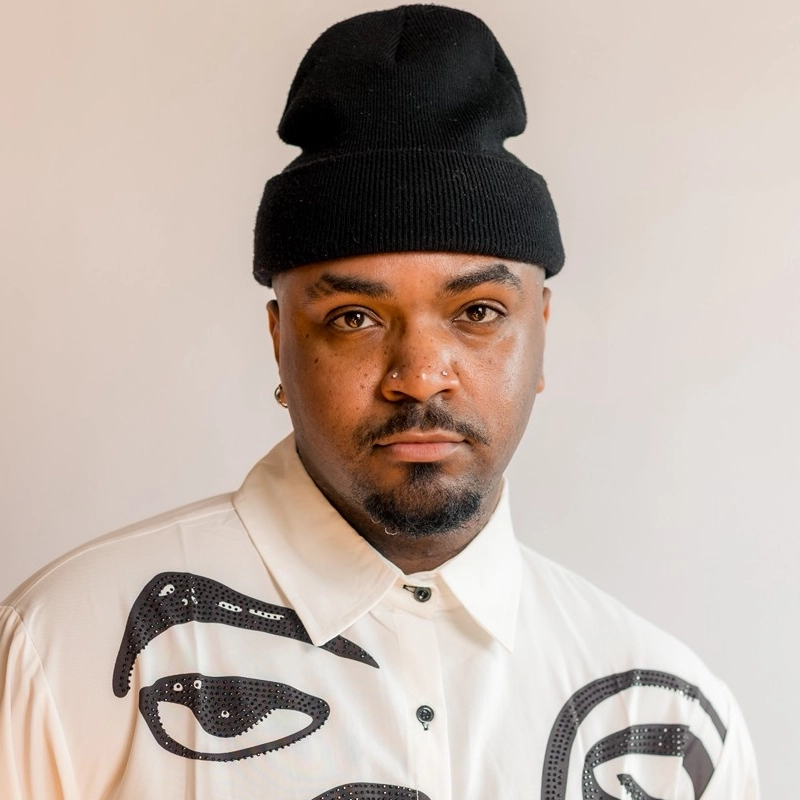 A man with a short beard and mustache wears a black beanie, a white button-up shirt with black abstract designs, and a nose ring. He has a neutral expression and is pictured against a plain, light-colored background.