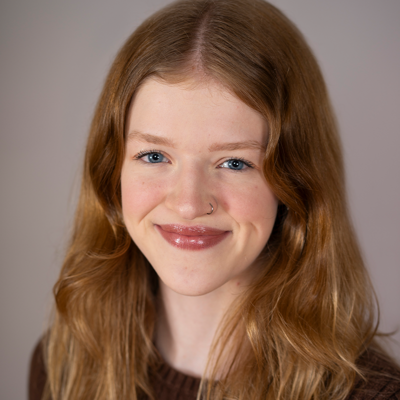 Chloee Brill, a young woman with long, wavy blonde hair, smiles at the camera. She has fair skin, blue eyes, and a nose ring. She wears a brown top and poses against a softly lit gray background.