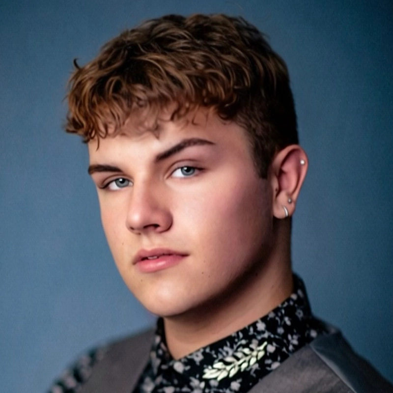Young person with short, curly light brown hair, blue eyes, and an earring in the left ear. Samantha Yakaitis wears a floral black shirt with a gray vest, posed against a blue background, looking confidently at the camera.