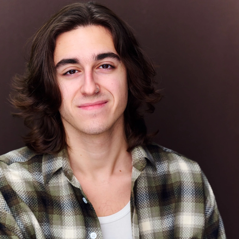 A young person with long brown hair, wearing a green and white plaid shirt over a white top, smiles gently against a plain dark background. They have a nose ring and a relaxed, confident expression—capturing the easygoing charm of Augustine Maiorino.