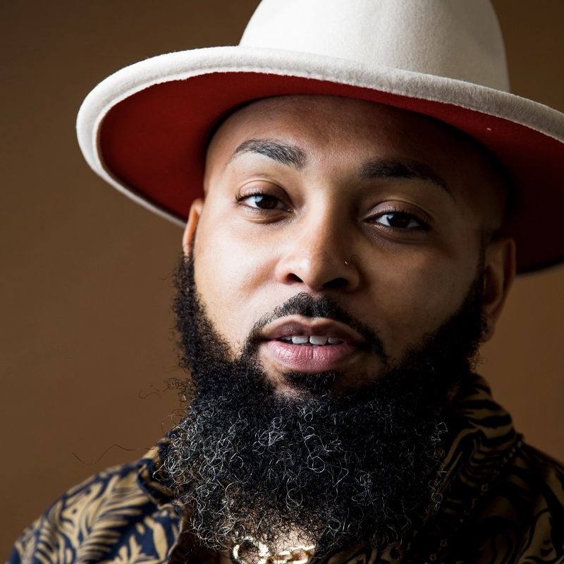Rashad McPherson, sporting a full, dark beard flecked with gray, dons a wide-brimmed cream hat with a red underside. With a nose ring and arched eyebrows, he poses in a patterned shirt against a plain brown background.