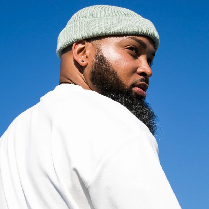A bearded man, resembling Rashad McPherson, wears a light green knit beanie and a white shirt as he looks over his shoulder. His skin glows in the sunlight against a clear blue sky, with a serious expression and a stud earring in his left ear.