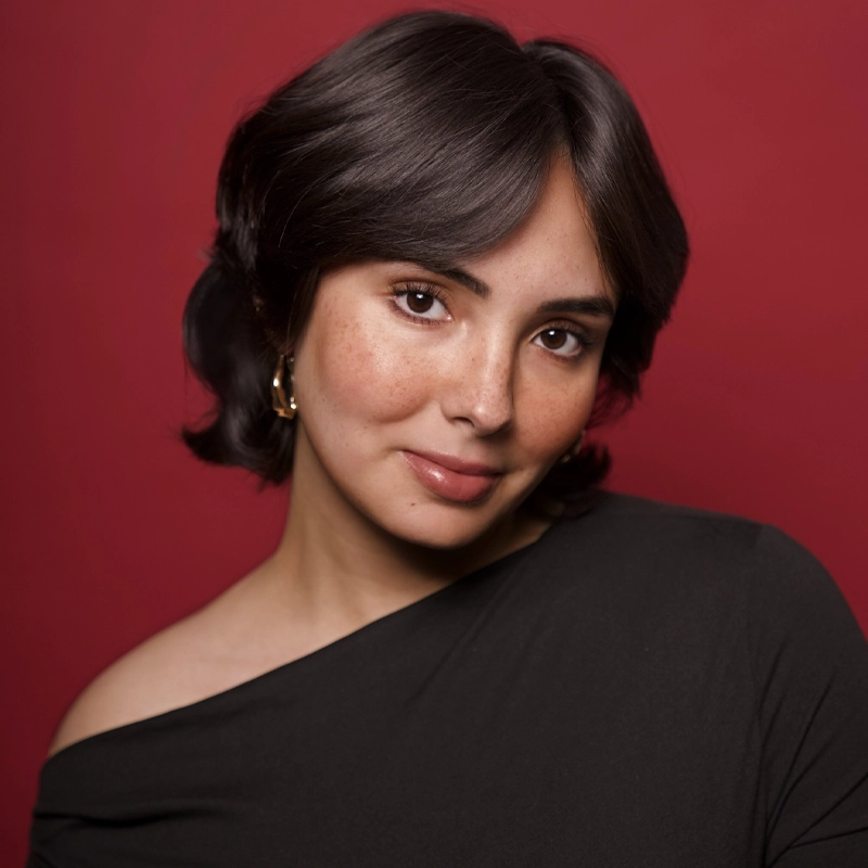 A young woman with short, dark hair and light brown skin poses against a deep red background. Sophia Orihuela wears a black, off-the-shoulder top and gold hoop earrings, her gentle smile and natural makeup highlighted by a slight tilt of her head.