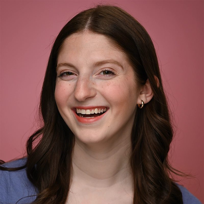 A young woman with long brown hair and fair skin smiles widely in front of a pink background. Samantha Yakaitis is wearing gold hoop earrings and a light blue top. Her expression is cheerful and friendly, and her makeup is natural.
