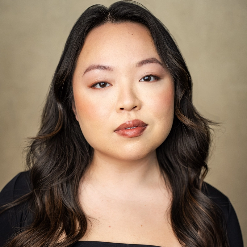 A woman with long, wavy dark hair and light skin looks at the camera with a neutral expression. She is wearing a black top and natural makeup with brown eyeshadow and glossy lips. The background is plain and light beige.