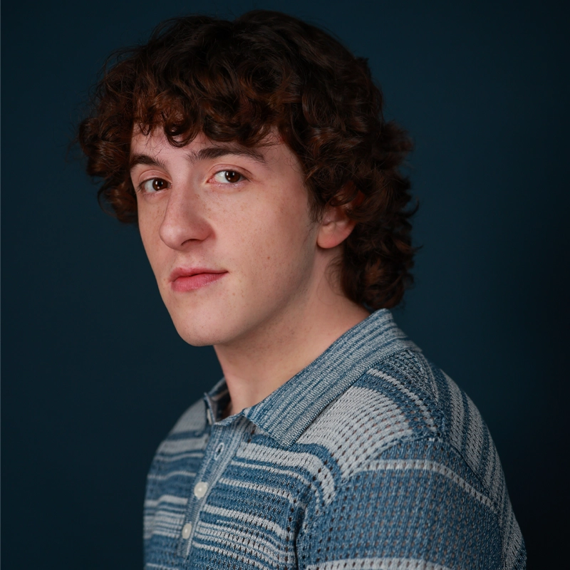 A portrait of Elijah Zurek, a young person with curly brown hair looking toward the camera, wearing a blue, textured knit polo shirt against a dark teal background.