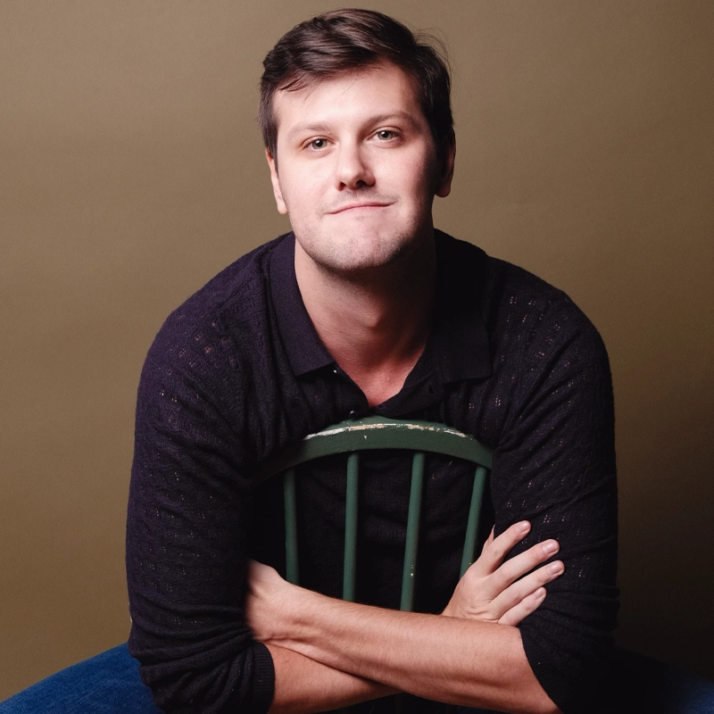 Noah Eisenberg, with short brown hair and a slight smile, sits backward on a green wooden chair, arms folded over the back. He wears a black long-sleeve shirt and blue jeans, set against a neutral brown background.