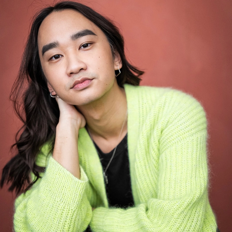 A person with long brown hair, wearing small hoop earrings and a fuzzy lime green cardigan over a black shirt, rests their head on their hand. They have a calm, thoughtful expression. The background is a solid reddish color.
