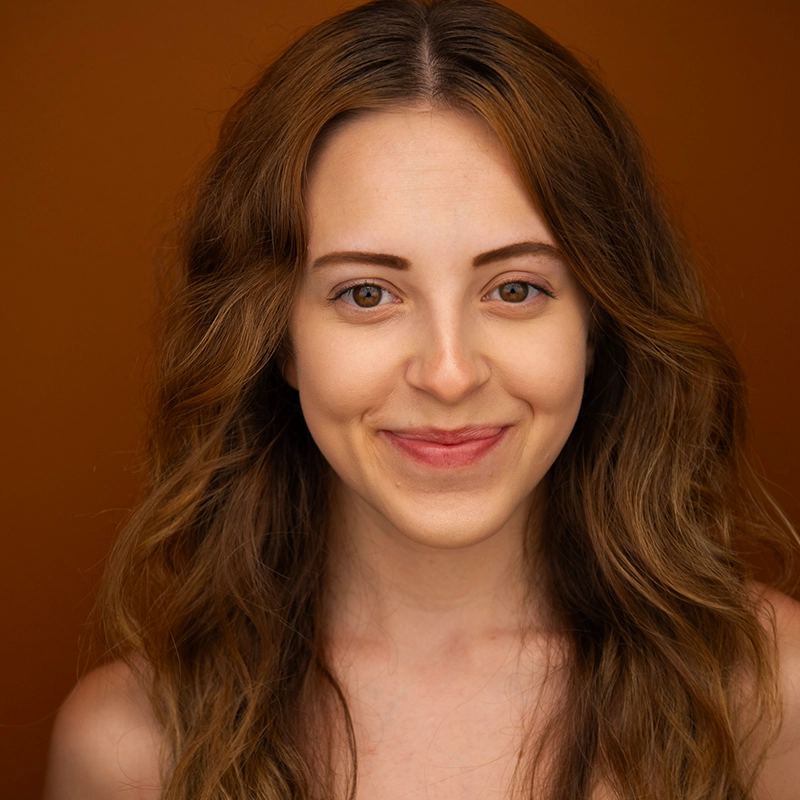 Allison Calabrese, a young woman with long, wavy, light brown hair, smiles gently. She has fair skin and faces the camera against a solid, warm orange background. Her makeup is minimal and natural, paired with a sleeveless top.